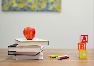 Various school items on a desk, including: a red apple fruit, four pyle books, colored pencils, and letter blocks.
