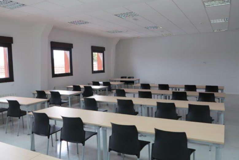 Black chairs with stylish back supports are neatly tucked in under rectangular desks facing the forward wall of a whitewashed square room purposed towards education as light filters through three square windows.