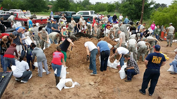 A large, multitude of people, some in casual clothing and others in military uniform, circling a dirt pile, use shovels to fill up white bags with the brown, rocky dirt while several pickup trucks parked in the background sit idle and green brush can be seen in the background.