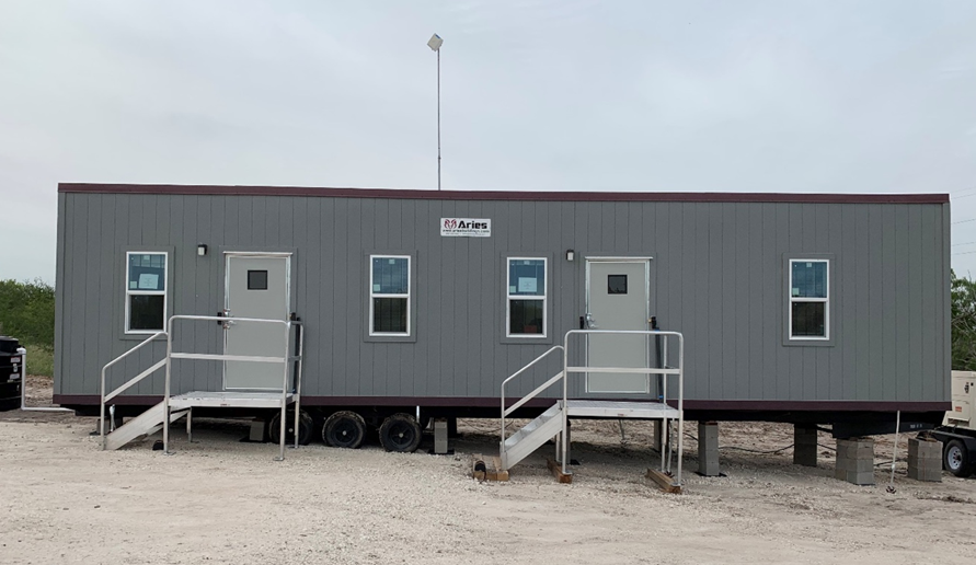 A long, grey trailer office, two doors and four windows, has two sets of metal stairs leading to the doors and sits connected to a supply of water, a long antenna on top, and an electric generator, all on a sandlot.