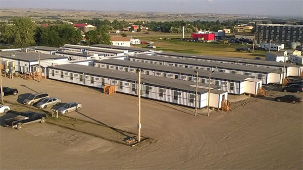 A wide panoramic view of a large, flat plain, brown in the distance, but near is a line of green trees and grass, in front of which are a four-story building and four long rows of modular trailers, white and grey, with wooden stairs at the ends and centers and a dirt parking lot with a handful of parked cars.
