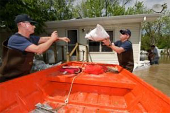 A small, off-yellow, one story house with a large square window and a screen door, has small green trees rising behind it and calm, light-brown water surrounding it while two men standing with water-proof overalls toss a sandbag to each other over a bright orange row-boat.