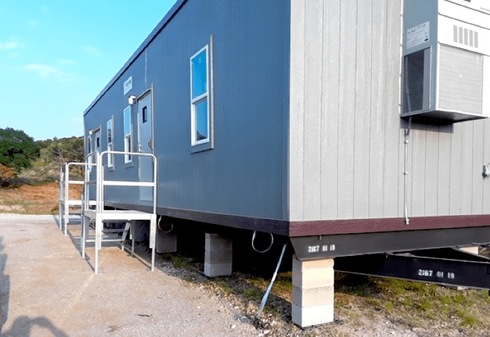 Two corner views of a grey trailer office on blocks that shows the metal ramps, air-conditioning unit, and placement on a grass and gravel lot on a blue sky day.