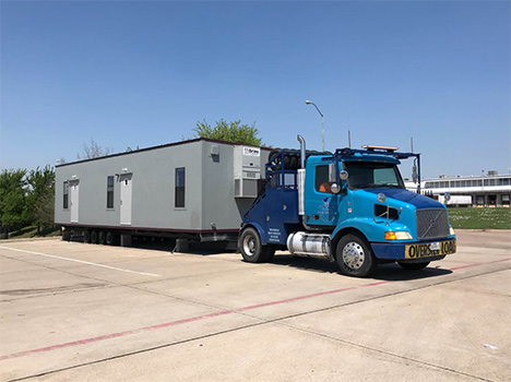 A bright blue truck transporting a grey pre-built modular building with maroon trim.