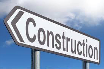 A close up of a white and black street sign in the form of an arrow pointing left in front of a blue sky with a puffy white cloud and with the word “Construction” printed in black letters.