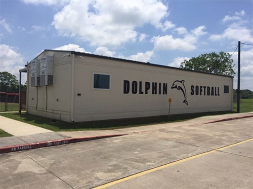 A long, off-white modular building with a Dolphins softball name and logo and two square windows at opposite ends stands quietly in front of trees and green grass on a calm, partly-cloudy day.