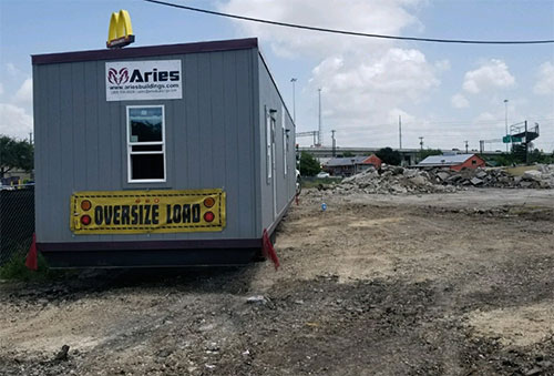 A rectangular, grey office building with a yellow “oversize load” sign and an Aries logo overlooks a construction site.