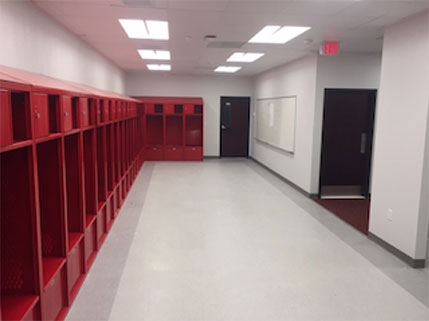 A long row of red sports lockers line one side of a spacious white room with fluorescentincandescent lighting and a whiteboard at the opposite wall of the lockers.
