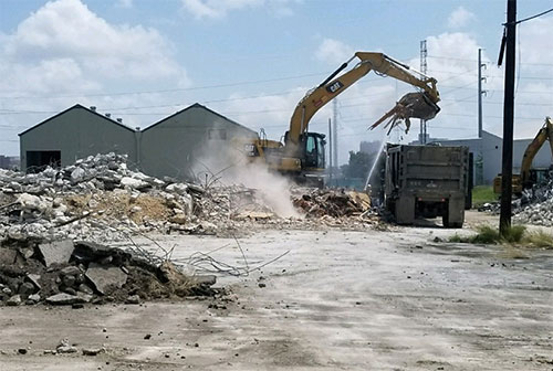 An excavator lifts its loader filled with rubble over a grey dump truck, blasting a dust cloud into the air.