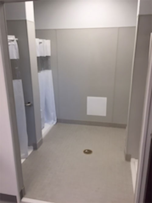 a grey and white shower room with white curtains for the showers and a central drain with a wide entrance door and a small square vent panel low on the far wall.