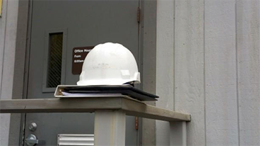 A white construction hardhat and binder perched on the corner of a wooden railing in front of a closed, grey door with a window, an office hours sign, a lever handle, mail slot, and a deadbolt lock.
