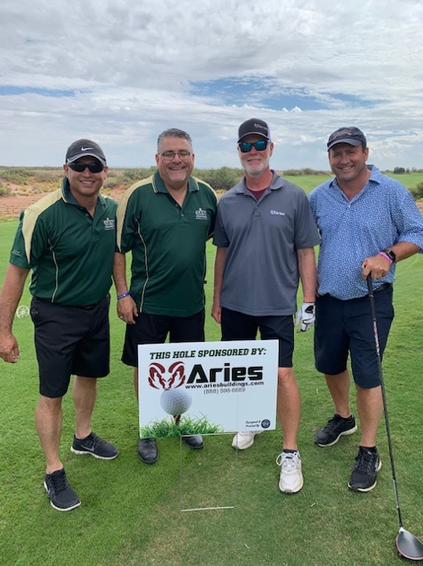 4 golfers gathered on a golf course on a warm sunny day around a knee-level sign that reads “This hole sponsored by Aries.”