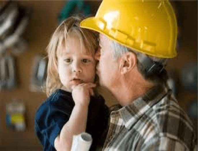 A man with a yellow hard hat and wearing a green, brown, and white plaid, flannel shirt is holding rolled-up paper, likely building plans, in one hand, and in the other he is holding and kissing a toddler wearing a navy colored top with strawberry blonde shoulder length hair and bangs.