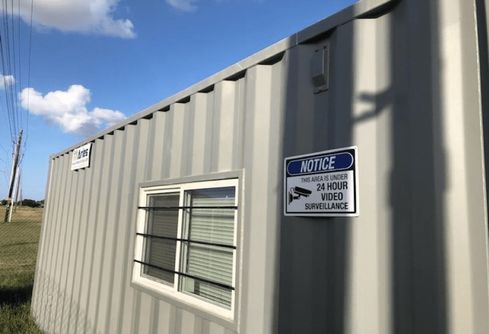 A grey, modular construction office exterior, with a window with bars over it, a sign that depicts a video camera on it and reads in all caps, “Notice: This area is under 24 hour video surveillance,” against the background of a blue sky, clouds, green grass, a utility pole, and a powerline.