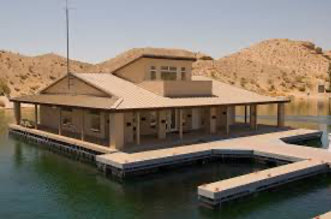 : A square, brown, modern wooden house with a wide, bonnet roof sitting on top of a large dock in the middle of a pond; in the background are a few small hills with sparse green vegetation.
