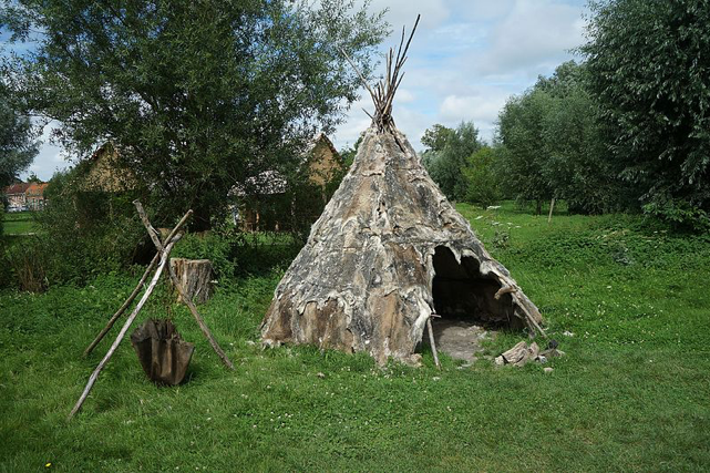 On verdant grass surrounded by trees sits a tee-pee made with thin branches, brown animal skins, and a small opening as a door. Through the trees you can see more tee-pees, resembling an early settlement. Attribution: Photograph by Pierre André Leclercq