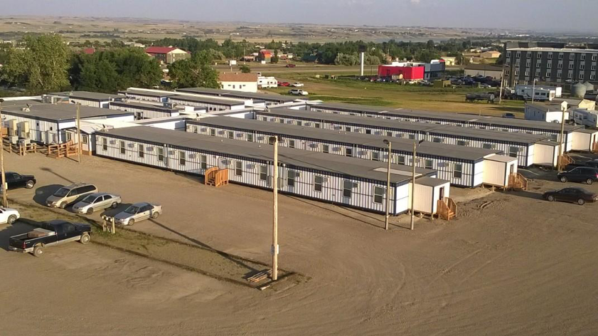 A wide panoramic view of a large, flat plain, brown in the distance, but near is a line of green trees and grass, in front of which are a four-story building and four long rows of modular trailers, white and grey, with wooden stairs at the ends and center and a dirt parking lot with a handful of parked cars.