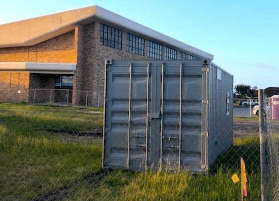 : In the foreground is a gray storage unit, on a patch of grass, surrounded by a fence; in the background is a large brown and red toned brick building with windows at the top.