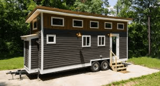 A gray, prefabricated tiny home on wheels on a concrete slab, with a hitch and several rectangular windows, a three-step staircase leading to the door, and green grass and trees in the background.