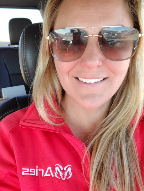 Katie Roman, VP of Workforce Housing at Aries seated in the drivers’ seat of a car; she’s a young attractive blonde woman with striking blue eyes, wearing a bright red Aries collared shirt.
