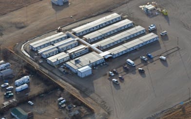 A bird’s eye view of an Aries service facility: several housing facilities lined up in rows of 4, with an adjoining hallway down the middle set on a stark landscape of brown dirt and with several cars in an adjacent parking lot.