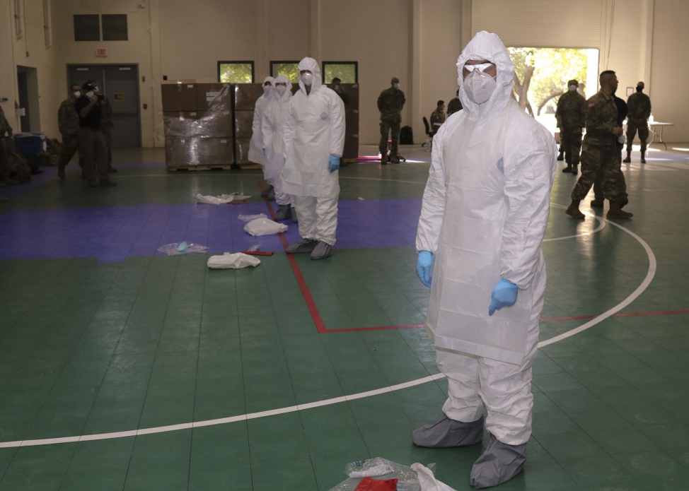 In a gym with green floor and the lines for a basketball court, a line of medics in white, protective medical suites with masks and protective glasses and grey covering for their shows look at the camera while military guards mill about around them. 