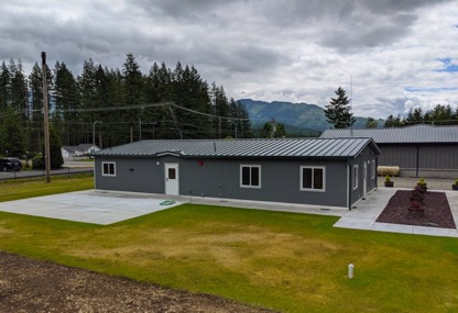 A panoramic shot of the exterior of two modular buildings, the nearest one with big square windows, a door on each side, a concrete patio, and a driveway. There is a yellow-green grass lawn and behind the buildings can be seen electric wires, pine trees, and small, round mountains. 