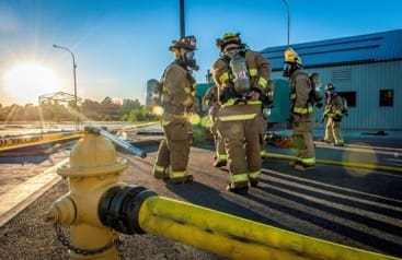 With a low, near horizon sun shining into the camera, the picture shows a close-up of a yellow fire hydrant with a thick, yellow hose attached, and a group of five firemen in the background in full gear and masks, casting long shadows and standing in front of a metallic building.