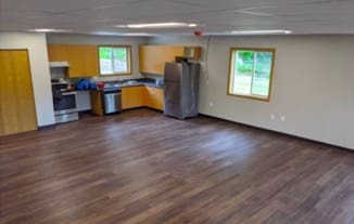 A large room with hardwood floors, square windows, and fluorescent lights is otherwise unfurnished. In one corner of the room is a kitchen area with a sink, dishwashing machine, refrigerator, oven, stove, and light orange cabinets.