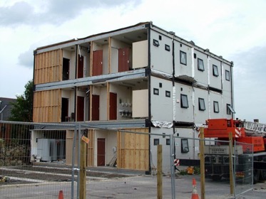 Behind a temporary metal fence, a three-story modular building is being assembled, the square modules are stacked on top of each other in 5 aligned rows, are covered in white wrap, and have one large window or two small windows in each section, while in front of the building an orange crane sits at rest. 