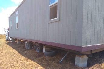 The view along a gray modular building with three vertical, rectangular windows. The trailer is still attached to a large truck and is being installed on concrete blocks. 