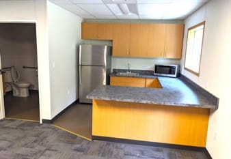 Interior picture of a kitchen next to an open bathroom. The kitchen has a wide, grey L-shaped counter, a microwave, a refrigerator, wooden cabinets, a sink, and a large window.