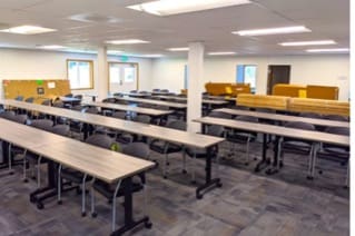 A large square room that looks like a classroom with two white columns in the middle, many fluorescent lights on the ceiling, a patterned carpet on the floor, double doors, wide windows, and rows of long tables and forward-facing seats.