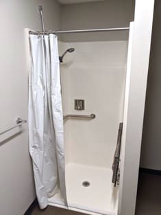 A full shower stall with a removable shower head, a grab bar, a drain in the middle of the floor or the enclosure, a white shower curtain, and a towel bar on the wall outside.