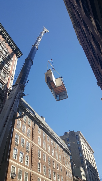 : Under a clear blue sky, a tall, narrow crane rises high into the sky, and the end is suspending a module for a building.