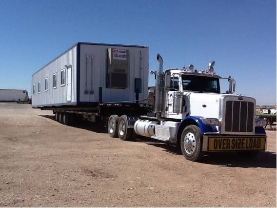 A large, white semi-truck labeled “oversize load” on the front bumper in a gravel lot has a large, rectangular mobile office on a trailer, and the office has 5 square windows along the side, a white rectangular door, and an air conditioning unit on the end.