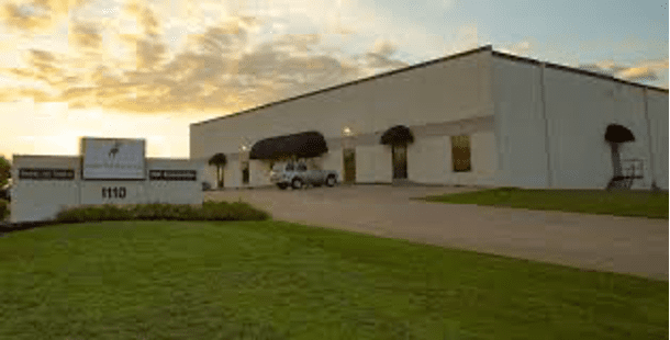 A low sun colors the sky and clouds a bright gold behind a large modular building with green awnings. A car is parked in front, and there is a sign that says “Aries,” on a green lawn.