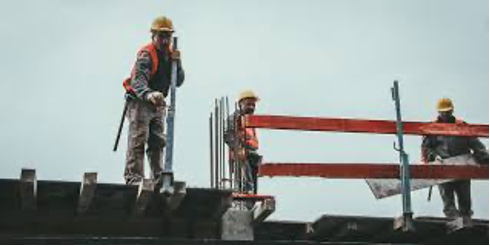Three construction workers wearing orange safety vests and yellow hard hats on top of a structure, one of the workers looks down as if speaking with someone standing below, while another moves a rectangular plank, and steel rods rise into the air above the platform of an unfinished building on a traditional construction site.