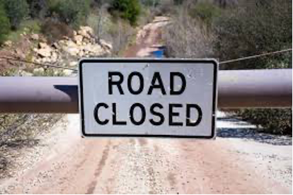 A dirt road is visible behind a close-up of a “Road Closed” sign attached to a horizontal, metal gate barring passage onto the road beyond.