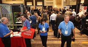 The busy World of Modular convention floor full of people milling around, some with lanyards around their necks, and many exhibition booths line both sides of a wide walkway with a tan and brown patterned carpet. 