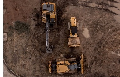 : A birds-eye-view shot of a leveler, a digger, and a bulldozer parked on a dirt lot with the tracks of their treads on the surrounding ground. 