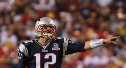 : Tom Brady, wearing a New England Patriots uniform, yells, and points towards his left while behind him is the blurry implication of a large crowd watching him play. 