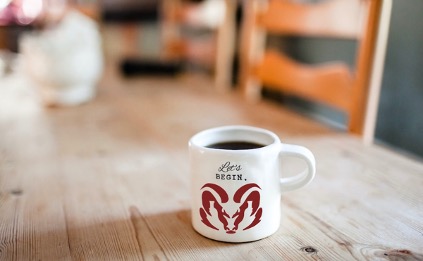 A coffee mug sits on a country-style wooden table as the morning sun streams in a nearby window. Written on the white coffee cup are the words, “Let’s begin,” above the official Aries’ burgundy ram-head logo.