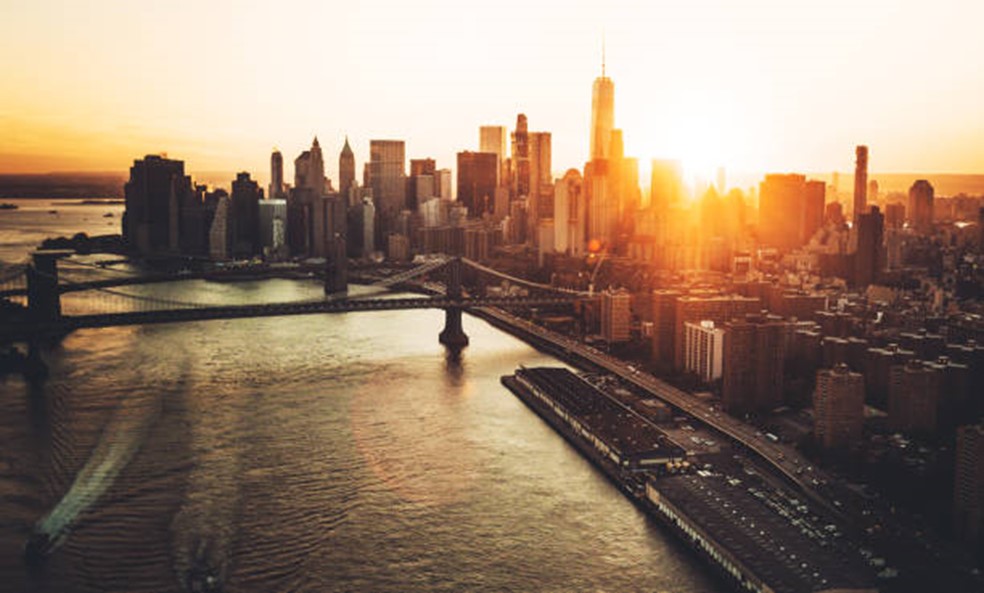 An aerial view over a river of the Manhattan skyline facing the sea and the sun on the horizon behind it.