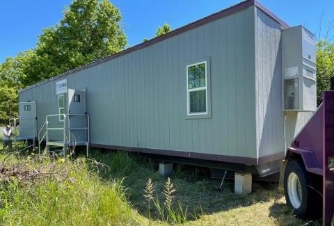 Picture of a grey modular office on a grassy lot hooked up to a purple truck that delivered the portable office to the spot, and the office has its two doors open and an air conditioning unit attached to the side.