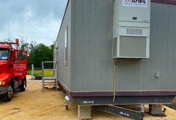 Picture of a modular building on blocks beside a red truck that delivered the mobile office to the brown gravel lot, and a metal staircase is installed next to the door of the trailer.