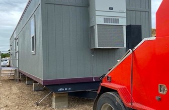 Picture of an ash gray modular building still connected to a dark orange delivery truck that placed the portable office on a rocky lot.