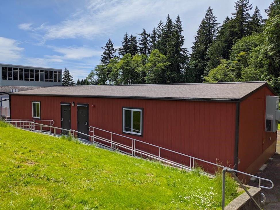 Behind a grassy knoll, a red portable classroom with two doors, ramps with handrails, and two white framed windows sits in front of the main school building and tall pine trees beneath a blue sky with wispy clouds.