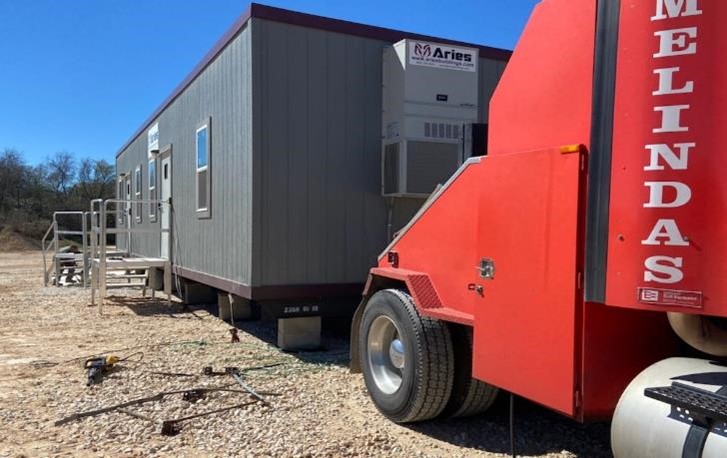 Pictured is a gray modular building still hooked up to a red delivery truck that has placed the portable office on a rocky lot under a blue sky.