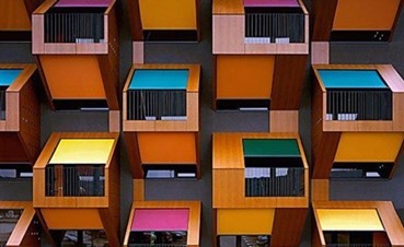 A bold and dynamic modular apartment design with rows of small, wooden boxes, each of which has a metal railing on the balcony in front of a large window. The tops of the boxed units are painted in striking shades of green, yellow, pink, and blue.